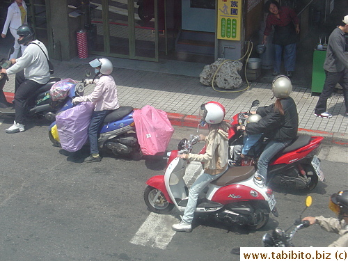 The woman in a pink shirt carries a lot of stuff in her scooter, and the lady behind her has a tiny passenger squeezed between her and the dashboard
